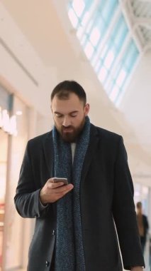 Modaya uygun bir adam telefonundaki güncellemeleri kontrol ederken hareketli bir alışveriş merkezinde geziniyor.