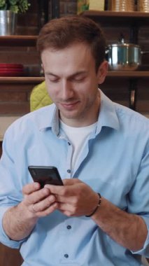 Çekici ve neşeli genç bir adamın portresi akıllı bir telefonla mutfak tezgahında oturuyor ve evdeki mesajları okuyor..