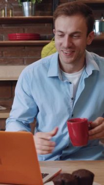 Mutfaktaki dizüstü bilgisayardan olumlu haberler okuyan başarılı girişimci gülümsüyor. Masada oturmuş, ekrana bakıp kahve içen gülümseyen genç adamın portresi..