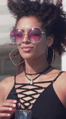 Güneş gözlüklü çekici genç bir kadın gülümsüyor gülüyor sahildeki bir partide kokteyl içtiği için çok ırklı gün batımını rahatlatıyor.