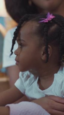 Bir kadın ve küçük bir kız kanepede rahat bir anı paylaşıyorlar. Pembe kurdeleli kız, neşe saçar kadın onu sevgiyle kucaklarken.