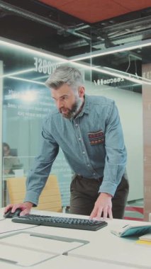 Sakallı bir adam, sanal gerçeklik kulaklığı takmadan önce masasındaki bir bilgisayara odaklanmış modern bir çalışma alanında teknolojiyle etkileşime geçer.