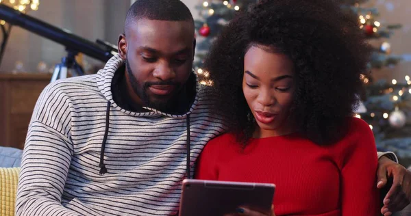 Tableti elinde tutan ve oyun oynayan genç bir kadın. Çekici Afrikalı çift kanepede oturuyor. Adam kız arkadaşına sarılıyor. Kapalı.