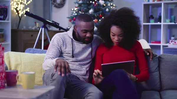 Sevimli genç Afrikalı aşık kanepede oturup internette makale okuyor. İnsanlar bir şey tartışıyor. İçeride. Süslü Noel ağacı arka planı