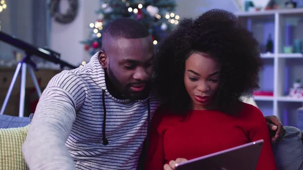 Genç Afrikalı çift kanepede oturup tablete bakıyor. İki insan birlikte vakit geçiriyor. Yakın plan. Kapalı.