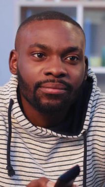 Spor kapüşonlu yakışıklı Afrikalı adam kanepede oturuyor ve güzel bir dairede televizyon izliyor. Adam televizyon kanalını değiştiriyor. Kapalı.