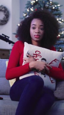 Kırmızı kazaklı güzel bir kadın kanepeye oturuyor ve süslü Noel ağacı ve teleskopunun arka planında yastığın arkasına saklanıyor. Kapalı.