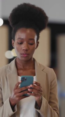 Şirketin içinde yürürken sosyal medyada akıllı telefonuyla mesaj yazan Afrikalı genç ofis yöneticisi kadın. İş ve teknoloji.