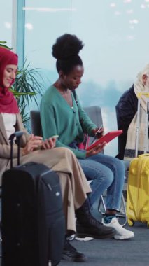 Bir grup gezgin, yoğun bir havaalanı bekleme alanında, uçuşlarını beklerken bavullarıyla birlikte dinleniyor ve cihazlarını çalıştırıyor.