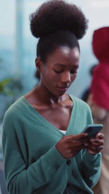 Çekici bir topuz içinde güzel siyah saçları olan ve rahat yeşil bir kazak giyen bir kadın dikkatle cep telefonuna bakıyor.