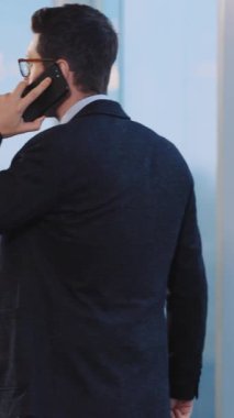 Takım elbiseli profesyonel bir adam cep telefonuyla konuşuyor, hem ayakta hem oturarak, hem de kendini işine adamış bir şekilde.