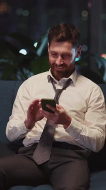 Genç Yetişkin İş Adamı Şık Ev Apartmanı 'nda koltukta oturuyor. Gün be gün Smartohone kullanıyor. Rahatlıyor..