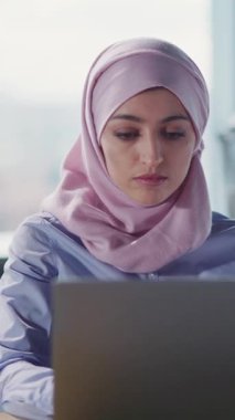 Kendini işine adamış ve tesettürlü genç bir kadın, çağdaş bir ofis alanında özenle dizüstü bilgisayarı üzerinde çalışıyor.