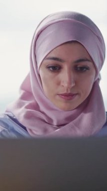 Pembe tesettürlü genç bir kadın, parlak ve ilham verici bir çalışma alanında çalışırken dizüstü bilgisayarına derinden odaklanır.