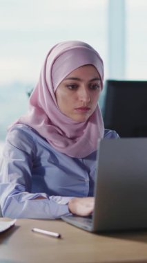 Pembe başörtüsü takan genç bir kadın, parlak bir ofis alanında dizüstü bilgisayarının çalışmasına derinden odaklanır.