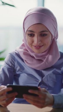 Pembe tesettürlü genç bir kadın modern ofis ortamında akıllı telefonunu kullanırken ışlı bir şekilde gülümsüyor.