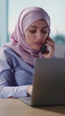 Şık bir başörtüsü takmış kararlı bir kadın modern bir çalışma alanında dizüstü bilgisayarı ve telefonuyla birden fazla iş yapıyor.