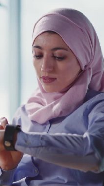 Modern ve çağdaş bir ofis ortamında, odaklanmış Müslüman bir kadın akıllı saatini güncellemeler için kontrol eder.