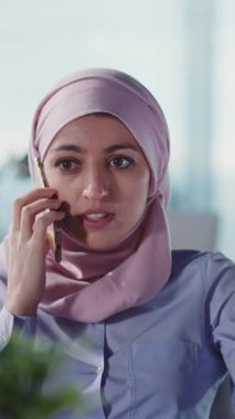 Çağdaş bir ofis ortamında, pembe tesettürlü odaklanmış genç bir kadın bir telefon konuşmasında yer alır.