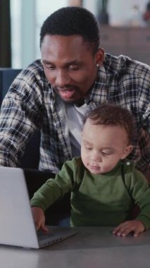 Afro-Amerikalı genç baba, oğlunun bilgisayar kullanmayı öğretmesine yardım ediyor. Oturma odasında birlikte oynuyorlar. Aileler. Çocuklar ve ebeveynler.