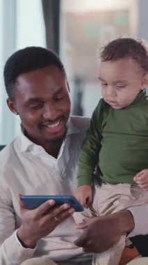 Afrikalı Amerikalı genç bir baba akıllı telefon kullanarak küçük bir çocuğu kucağına alıyor ve evde yeni şeyler öğretiyor. Kilitlenme. Yetiştirme.