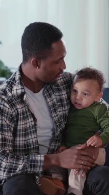 Afro-amerikan yakışıklı, mutlu, şişko, bebek ağlayan çocuğu kucağında tutuyor, birlikte oturma odasında oynaşıyor. Aile fotoğrafı..