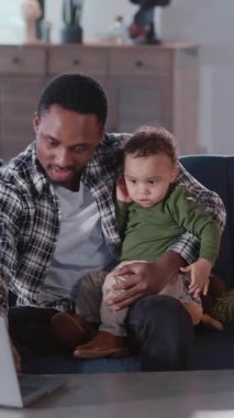 Meşgul genç Afrikalı baba dizüstü bilgisayarda bağımsız çalışıyor ve aynı anda evde küçük bir çocuğa bakıyor. Modern yaşam tarzı.