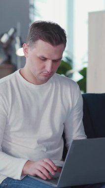 Genç ve çekici bir girişimci, beyaz bir adam. Dizüstü bilgisayarını kullanıyor. Ayrı oturma odasında otururken kameraya bakıyor. Serbest Çalışan Portresi.