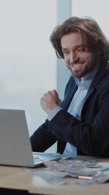 Sivri takım elbiseli odaklanmış bir işadamı, dizüstü bilgisayarına dalmış, modern bir ofise kendini adamış ve profesyonelliği somutlaştırmış bir şekilde bir masada oturuyor.