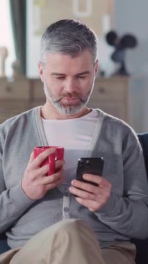 Oturma odasında cep telefonu kullanan yakışıklı milenyum çocuğu. İş adamı yetişkin mesajlaşması sosyal medyada sabahları kahve içiyor.