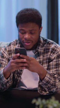 Genç bir adam, rahat bir ev ortamında modern dijital etkileşimi somutlaştıran akıllı telefonuna odaklanır.