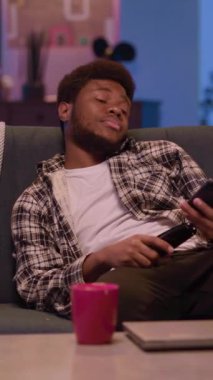 Genç bir insan elinde uzaktan kumandayla rahat bir şekilde kanepede uzanıyor, evinde sıcak bir akşamın tadını çıkarıyor.