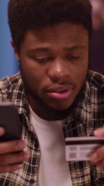 Genç bir adam elinde kredi kartıyla internet üzerinden alışveriş yaparken akıllı telefonuna odaklanmış görünüyor..