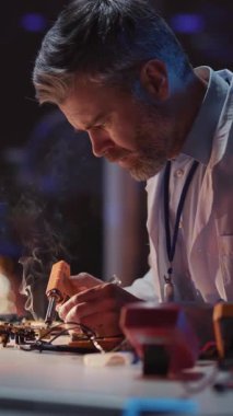 Beyaz önlüklü, sakallı yetişkin bir mühendis masa başında oturmuş İHA cihazını tamir ediyor. Teknik onarım, araştırma ofisi. Mühendislik.