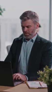 Gösterişli takım elbiseli profesyonel bir adam masasında oturuyor, dizüstü bilgisayarına odaklanıyor, kendine güveni, şık bir sakalı var, modern profesyonelliği sergiliyor.