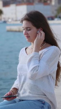 Çekici, genç, beyaz bir kadın deniz kenarındaki kumsalda oturmuş kulaklık takarak akıllı telefondan müzik dinliyor. Tatil konsepti. Boş zaman aktivitesi.