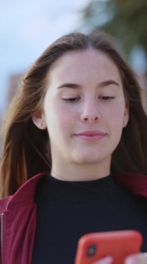 Tasasız, neşeli, beyaz bir kadın cep telefonu kullanıyor. Uygulamayı sosyalleştiriyor, mesajlaşıyor, saçını dışarıda okşuyor..