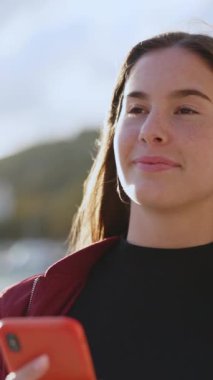 Uzun saçlı, kumsalda cep telefonu uygulaması kullanan güzel ve çekici bir kadın açık havada güneşli bir havada yürüyor..