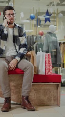 Meşgul bir alışveriş merkezindeki bankta oturan genç bir adam, telefonuyla konuşuyor, ona bakarken, canlı müşteriler ve mağazalar arasında yansıtıyor.