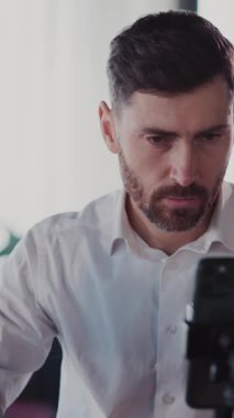 Bu video, sakallı bir adamın günlük rutinini gösteriyor. Önce telefonuna odaklanıyor, sonra kahvenin tadını çıkarıyor ve bilgisayarına bağlanıyor.