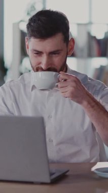 Neşeli bir adam masada rahatlar, kahvesini yudumlar ve laptopuyla çalışır, beyaz bir gömlek giyer, pozitif enerji ve verimlilik yayar.