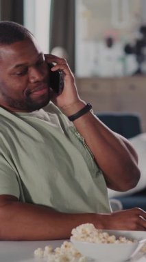 Bir adam cep telefonuyla sohbet ederken tesadüfen masada patlamış mısırdan zevk alır, rahat bir mutfakta huzur verici çoklu görevler yapar.