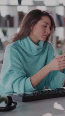 Rahat bir ev ofisinde, mavi kazaklı bir kadın masasında, klavyeyle müzik çalıyor ve telefonuna bağlanıyor.