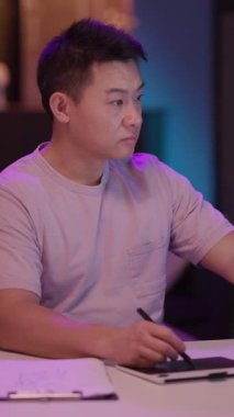 Mor gömlekli bir adam masada oturuyor, not yazarken bilgi için tablet kullanıyor. Sahne, onun teknoloji ve geleneksel araçları harmanlamaya odaklandığını vurguluyor.