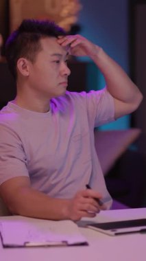 Bir adam eli alnında, derin düşüncelere dalmış bir masada oturuyor. Daha sonra bir tablete odaklanır, bir stil ile yazma veya çizim yapar.