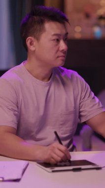 Mor gömlekli bir adam bir masada oturup tablete yazmaya odaklanmış. Ara sıra yukarı bakar, izleyiciyle minimalist bir ortamda bağlantı kurar.