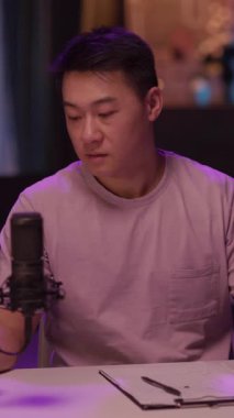 Mor gömlekli bir adam, bir masada mikrofonla oturmuş, düşüncelerini paylaşmaya hazırlanırken pembe bardaktan yudumluyor.