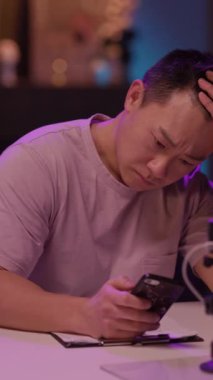 Sessiz bir odada, mor gömlekli bir adam masada oturuyor, mikrofonla derin düşüncelere dalmış, duygusal yansımayı ima ediyor.