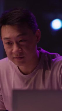 Pembe gömlekli bir adam loş bir odadaki bir masaya odaklanır ve samimi bir kayıt için mikrofona konuşmaya hazırlanırken laptopuyla meşgul olur.