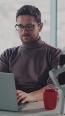 Gözlüklü bir adam bir masada oturuyor, özenle dizüstü bilgisayarı üzerinde çalışıyor, cihazın çeşitli görevleriyle çok yakından ilgileniyor.